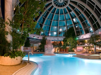 Große Glaskuppel der Westfalen-Therme am Abend, beleuchtetes Innenbecken mit Pflanzen und Liegen.