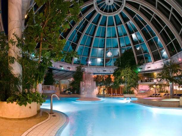 Große Glaskuppel der Westfalen-Therme am Abend, beleuchtetes Innenbecken mit Pflanzen und Liegen.