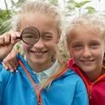 Zwei lächelnde Kinder in bunten Jacken, eines mit einer Lupe vor dem Auge, stehen im Grünen.