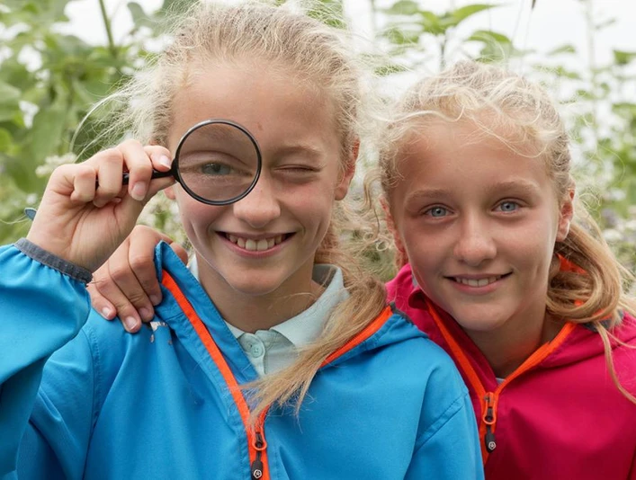 Zwei lächelnde Kinder in bunten Jacken, eines mit einer Lupe vor dem Auge, stehen im Grünen.