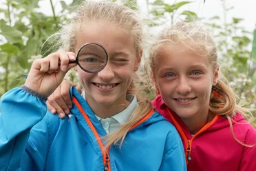 Naturentdecker Zwei lächelnde Kinder in bunten Jacken, eines mit einer Lupe vor dem Auge, stehen im Grünen.