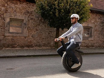 Mann fährt auf einem elektrischen Einrad, trägt einen Helm und Sonnenbrille, vor einer Steinfassade.