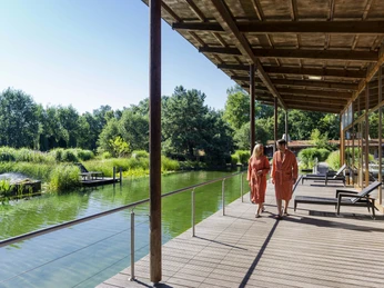 Zwei Personen spazieren auf einem Holzsteg am Badesee Järvesauna, umgeben von üppiger Natur.