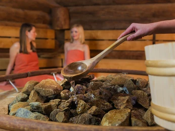 Zwei Frauen sitzen in einer Saunakammer, während Wasser mit einem Holzlöffel auf heiße Steine gegossen wird.