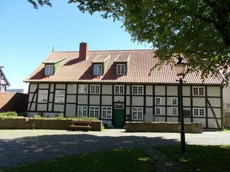 Fachwerkhaus mit rotem Ziegeldach, grüner Tür, umgeben von Bäumen und gepflastertem Platz.