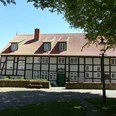 Bürgerhaus Preußisch Oldendorf Fachwerkhaus mit rotem Ziegeldach, grüner Tür, umgeben von Bäumen und gepflastertem Platz.
