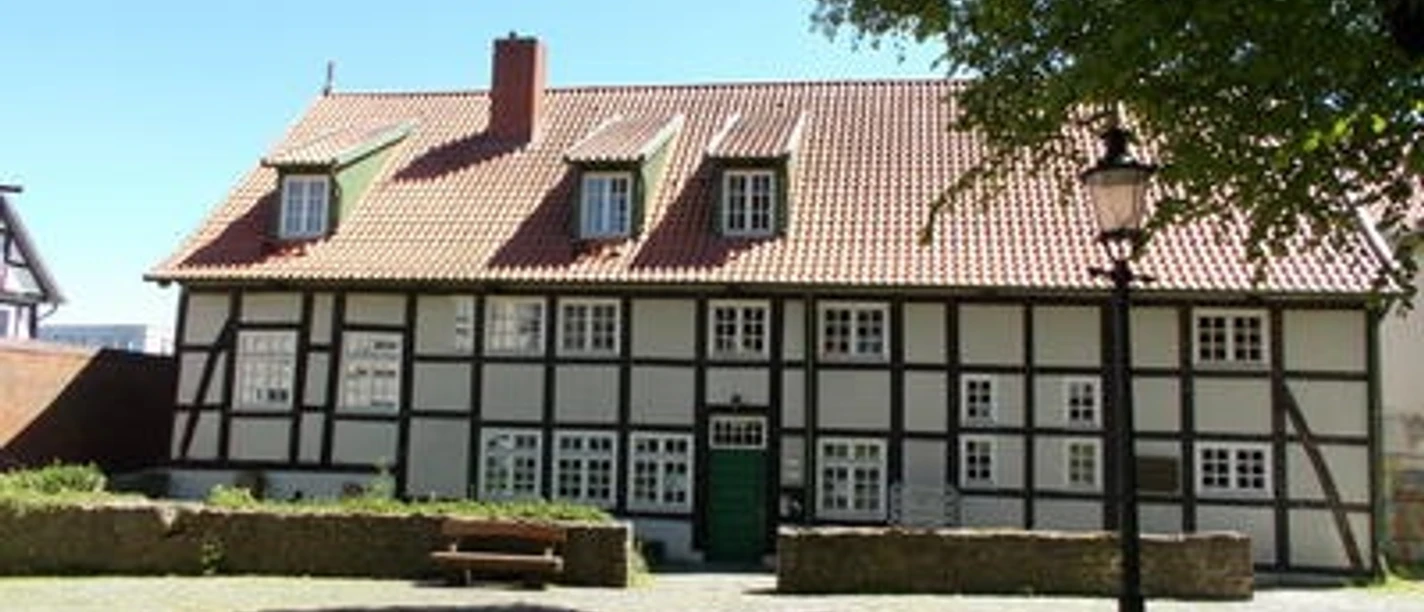 Bürgerhaus Preußisch Oldendorf Fachwerkhaus mit rotem Ziegeldach, grüner Tür, umgeben von Bäumen und gepflastertem Platz.