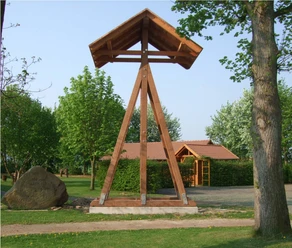 Glocke am historischen Dorfplatz Getmold Holzkonstruktion trägt Glocke auf Dorfplatz, umgeben von Bäumen und einem kleinen Gartenhaus.