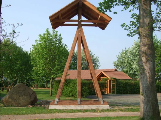 Glocke am historischen Dorfplatz Getmold Holzkonstruktion trägt Glocke auf Dorfplatz, umgeben von Bäumen und einem kleinen Gartenhaus.
