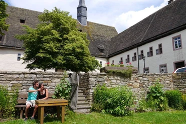 Garten in der Klosteranlage Ein Klostergarten mit alten Steinmauern, einem Holztisch und zwei entspannenden Personen auf einer Bank.