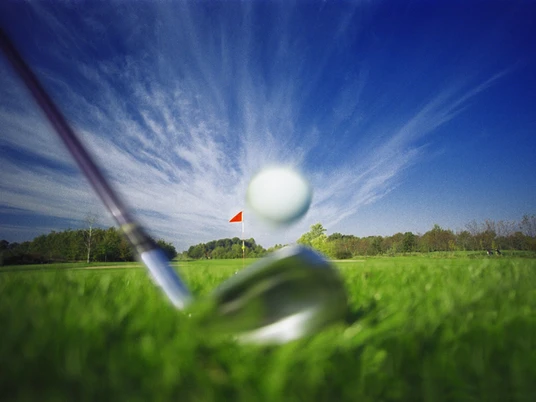 Golfclub Marienfeld Schläger mit Ball Ein Golfschläger beim Schlag trifft nahe eines Lochs mit roter Fahne auf einem grünen Golfplatz.