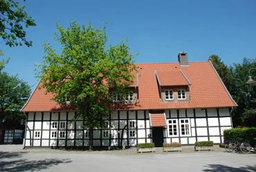 Fachwerkhaus mit rotem Ziegeldach, umgeben von Bäumen und blühendem Garten, an einem sonnigen Tag.