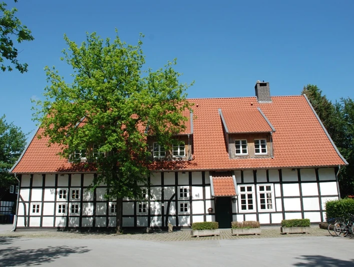 Fachwerkhaus mit rotem Ziegeldach, umgeben von Bäumen und blühendem Garten, an einem sonnigen Tag.