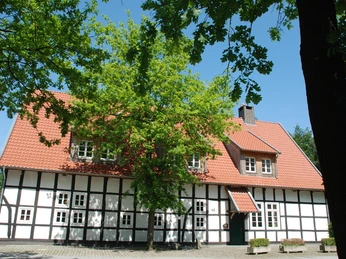 Fachwerkhaus mit roten Dachziegeln und weißen Wänden umgeben von grünen Bäumen unter blauem Himmel.