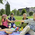 Eine Familie genießt bei Sonnenschein ein Picknick auf einer Wiese vor einem historischen Schloss.