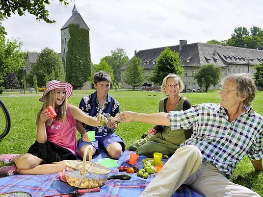 Eine Familie genießt bei Sonnenschein ein Picknick auf einer Wiese vor einem historischen Schloss.