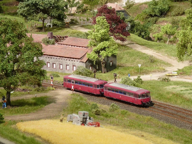 Modellbundesbahn Schienenbus im Weserbergland Ein roter Schienenbus passiert eine ländliche Modelllandschaft mit einem Fachwerkgebäude und Feldern.