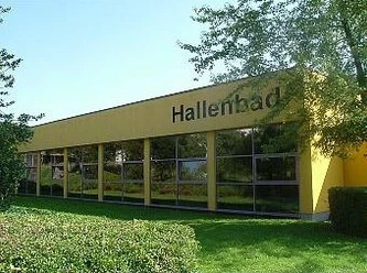 Gelbes Gebäude eines städtischen Hallenbades mit großen Fenstern, umgeben von grüner Landschaft.