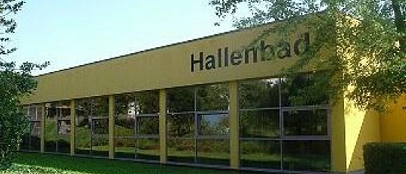 Städtisches Hallenbad Gelbes Gebäude eines städtischen Hallenbades mit großen Fenstern, umgeben von grüner Landschaft.