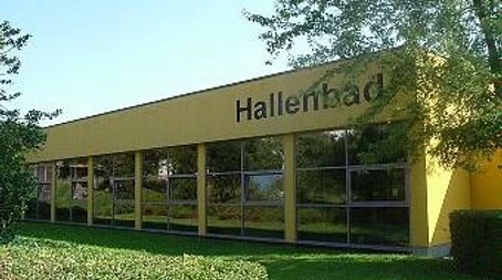 Städtisches Hallenbad Gelbes Gebäude eines städtischen Hallenbades mit großen Fenstern, umgeben von grüner Landschaft.