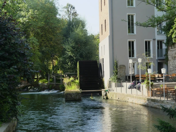 Historische Mühle mit Wasserrad, umgeben von Grün, angrenzend die Terrasse eines einladenden Cafés.