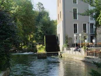 Stümpelsche Mühle und Terrasse des Mühlencafés Historische Mühle mit Wasserrad, umgeben von Grün, angrenzend die Terrasse eines einladenden Cafés.