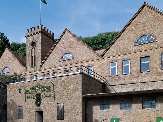 Historisches Backsteingebäude mit Zinnengiebel und Brauereilogos, umgeben von bewaldeter Landschaft.