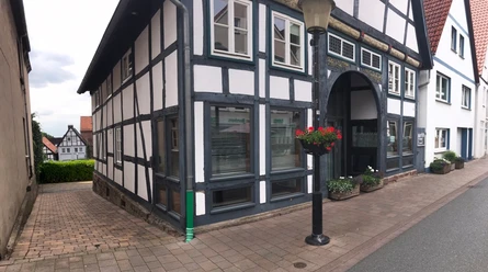 HausParkplatz_o.A.jpg Fachwerkhaus an einer gepflasterten Straße mit Blumenampel und klassischer schwarz-weißer Fassadengestaltung.