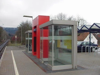 Bahnhof Sandebeck Moderner Bahnhof Sandebeck mit roten Schutzunterständen, Gleisen und umliegendem bewaldeten Hügel.