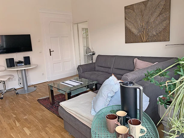 Wohnzimmer Apartment Gerome Helles Wohnzimmer mit grauem Sofa, Glastisch und Pflanzen. Moderne Einrichtung mit TV an der Wand.