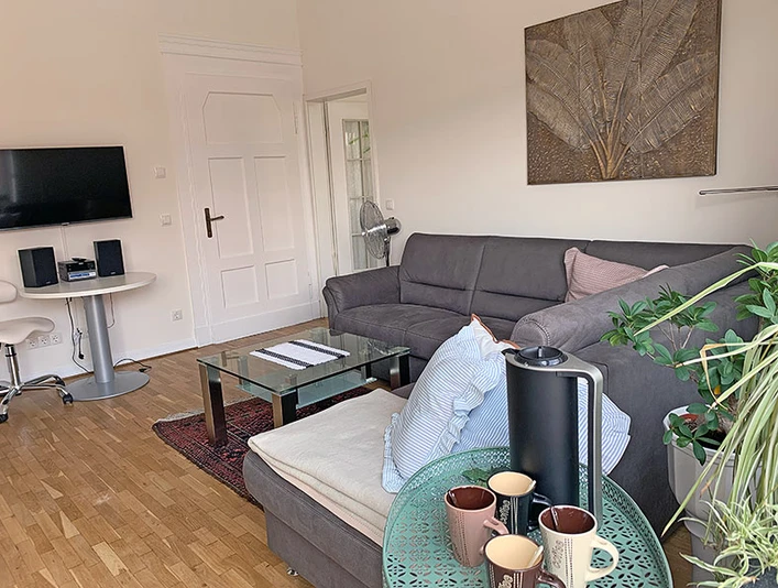 Wohnzimmer Apartment Gerome Helles Wohnzimmer mit grauem Sofa, Glastisch und Pflanzen. Moderne Einrichtung mit TV an der Wand.