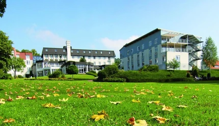 Große Grünfläche mit herbstlichen Blättern, dahinter zwei moderne Gebäude der Klinik Dreizehnlinden.