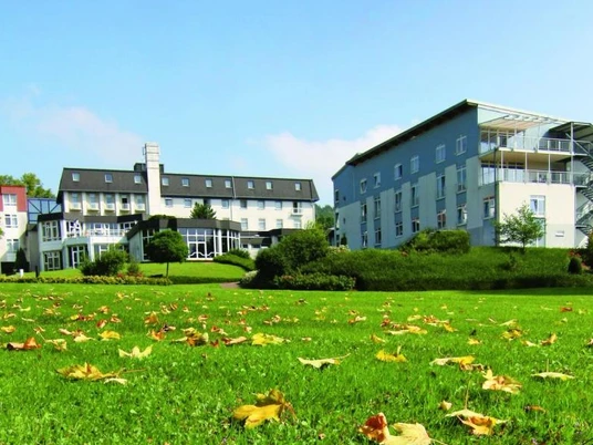 Klinik Dreizehnlinden Große Grünfläche mit herbstlichen Blättern, dahinter zwei moderne Gebäude der Klinik Dreizehnlinden.