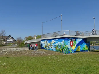 Graffiti am ehemaligen Kraftwerk bei der Fußgängerbrücke Graffiti in leuchtenden Farben ziert die Wand unter einer Fußgängerbrücke, umgeben von Grünflächen.