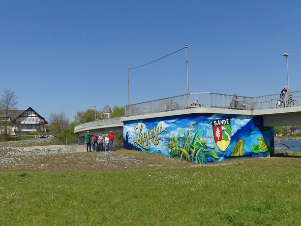 Graffiti am ehemaligen Kraftwerk bei der Fußgängerbrücke Graffiti in leuchtenden Farben ziert die Wand unter einer Fußgängerbrücke, umgeben von Grünflächen.