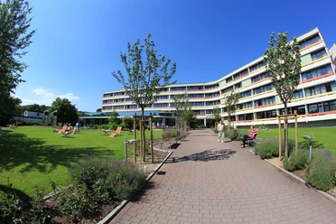 Moderner Klinikbau mit gepflegter Gartenanlage, Sonnenschirmen und Liegestühlen, klarer Himmel.