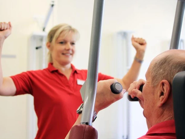 Frau in rotem Polo-Hemd leitet älteren Mann beim Krafttraining an einer Fitnessmaschine an.
