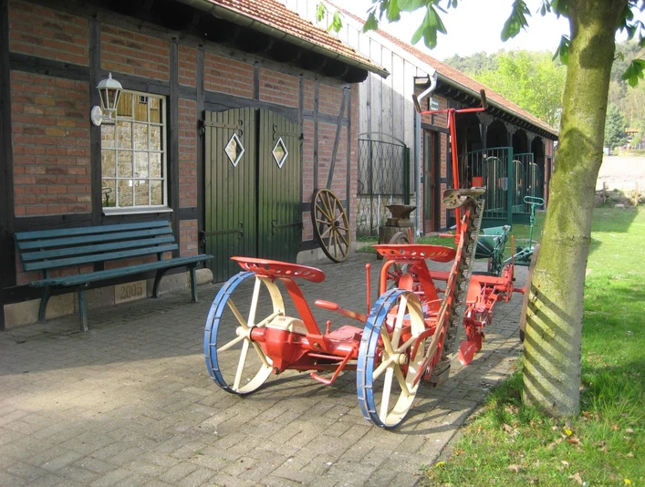 Landmaschinenmuseum Hörstel Rote, antike Pflugmaschine auf gepflastertem Hof vor einem Fachwerkgebäude mit grünen Türen.