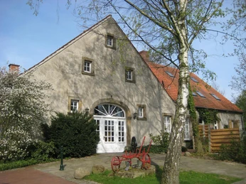 Landmaschinenmuseum Hörstel Historischer Bauernhof mit rotem Pflug im Vordergrund, umgeben von Bäumen und blühenden Sträuchern.