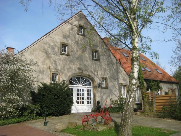 Landmaschinenmuseum Hörstel Historischer Bauernhof mit rotem Pflug im Vordergrund, umgeben von Bäumen und blühenden Sträuchern.