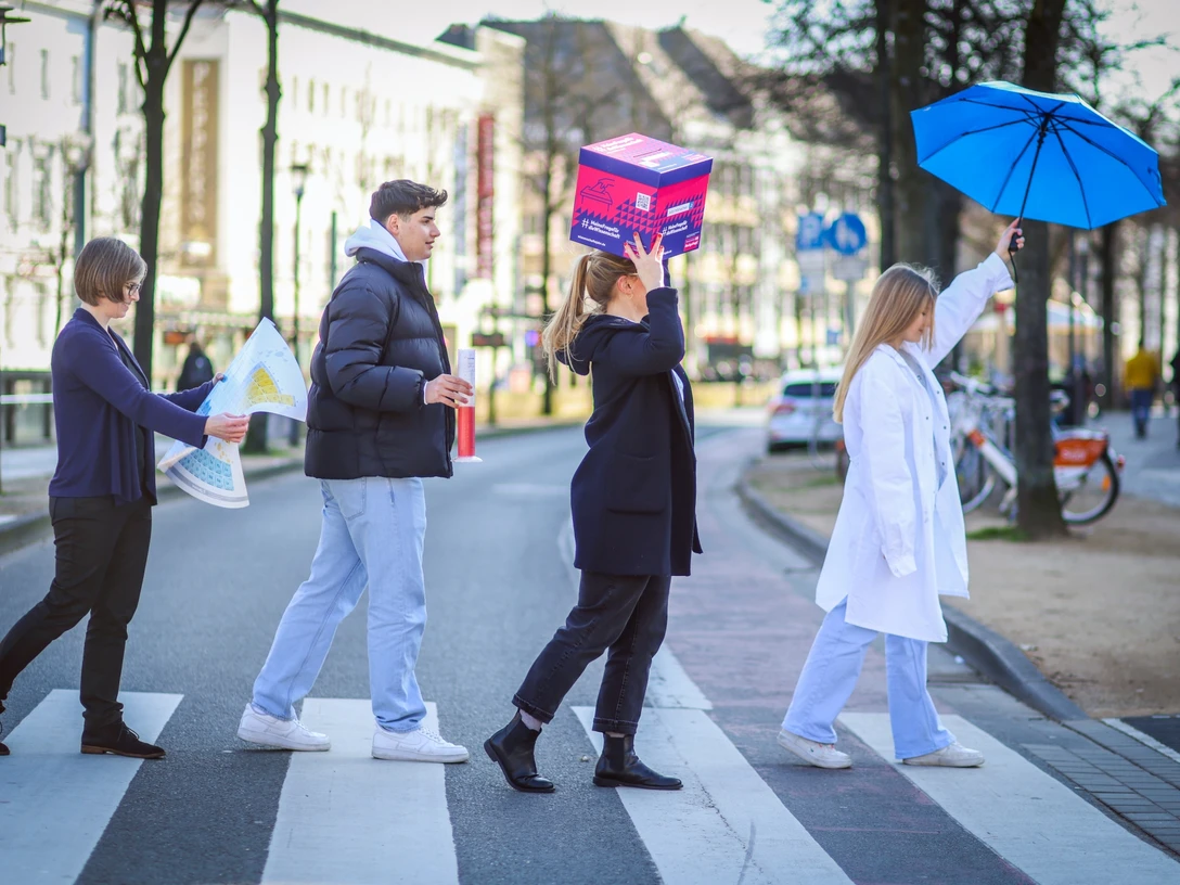 Science-Seeing-Tour durch Bielefeld.jpg Vier Menschen überqueren einen Zebrastreifen, zwei mit ungewöhnlichen Accessoires: Schachtel und Regenschirm.