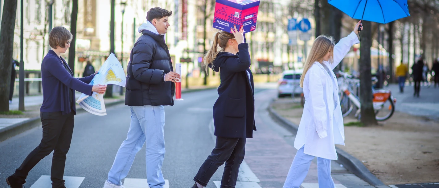 Science-Seeing-Tour durch Bielefeld.jpg Vier Menschen überqueren einen Zebrastreifen, zwei mit ungewöhnlichen Accessoires: Schachtel und Regenschirm.