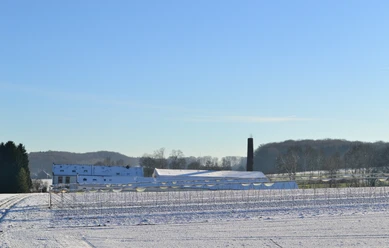 Blick auf die ehemalige Knochenleimfabrik Schneebedeckte Felder mit bäuerlichen Gebäuden und einem Schornstein. Winterlandschaft unter blauem Himmel.