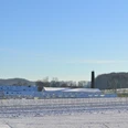 Blick auf die ehemalige Knochenleimfabrik Schneebedeckte Felder mit bäuerlichen Gebäuden und einem Schornstein. Winterlandschaft unter blauem Himmel.