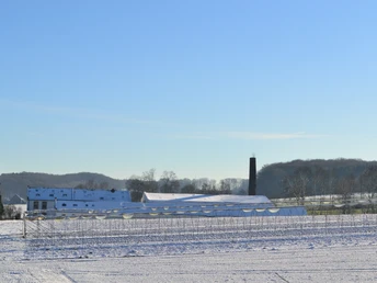 Blick auf die ehemalige Knochenleimfabrik Schneebedeckte Felder mit bäuerlichen Gebäuden und einem Schornstein. Winterlandschaft unter blauem Himmel.