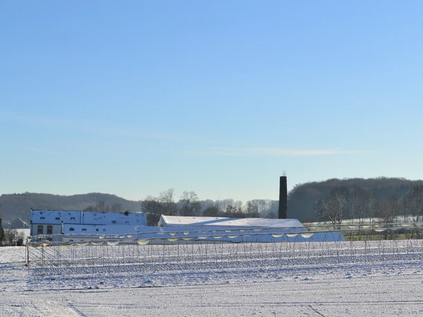 Blick auf die ehemalige Knochenleimfabrik Schneebedeckte Felder mit bäuerlichen Gebäuden und einem Schornstein. Winterlandschaft unter blauem Himmel.