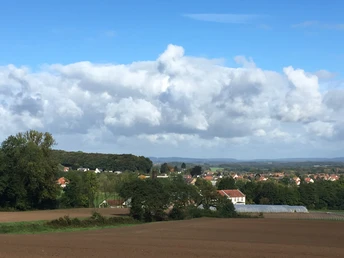 Weitläufige Landschaft mit Feldern, Wald und einer kleinen Ortschaft unter blauem Himmel mit Wolken.