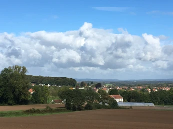 Weitläufige Landschaft mit Feldern, Wald und einer kleinen Ortschaft unter blauem Himmel mit Wolken.