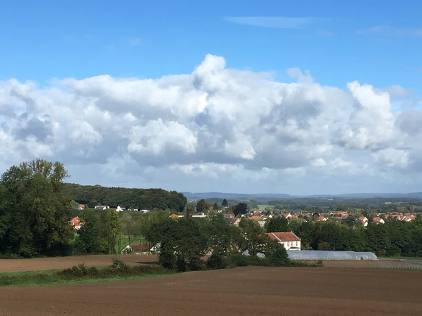 Blick auf Werther (Westf.) Weitläufige Landschaft mit Feldern, Wald und einer kleinen Ortschaft unter blauem Himmel mit Wolken.