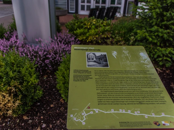 Informationsschild mit Text und Abbildungen über die Bauernopfergruppe aus Arrode, umgeben von Pflanzen.